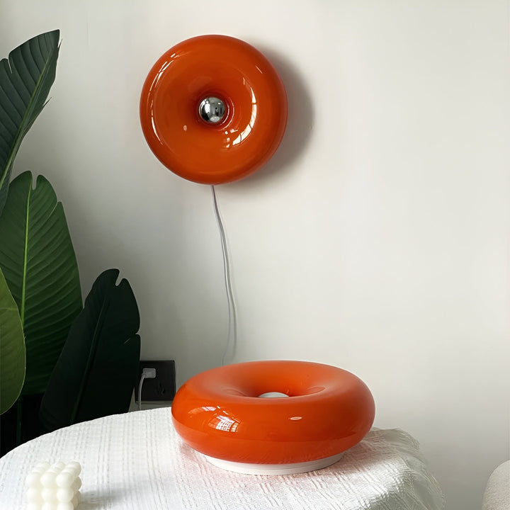 Orange donut-shaped LED lamp for table or wall, made of glass and metal, offering warm glow and whimsical décor touch.